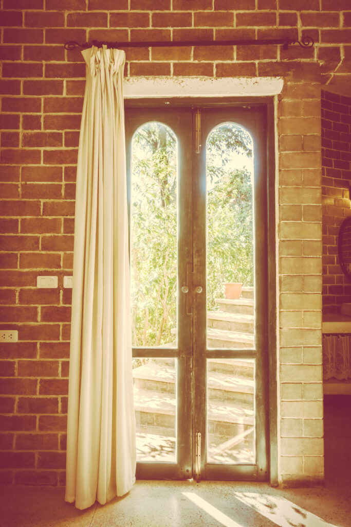 Rideau isolant long de couleur beige clair ou lin sur porte-fenêtre cintrée en bois foncé. Mur de briques apparentes rustiques. Décoration vintage ou style café européen laissant passer la lumière.