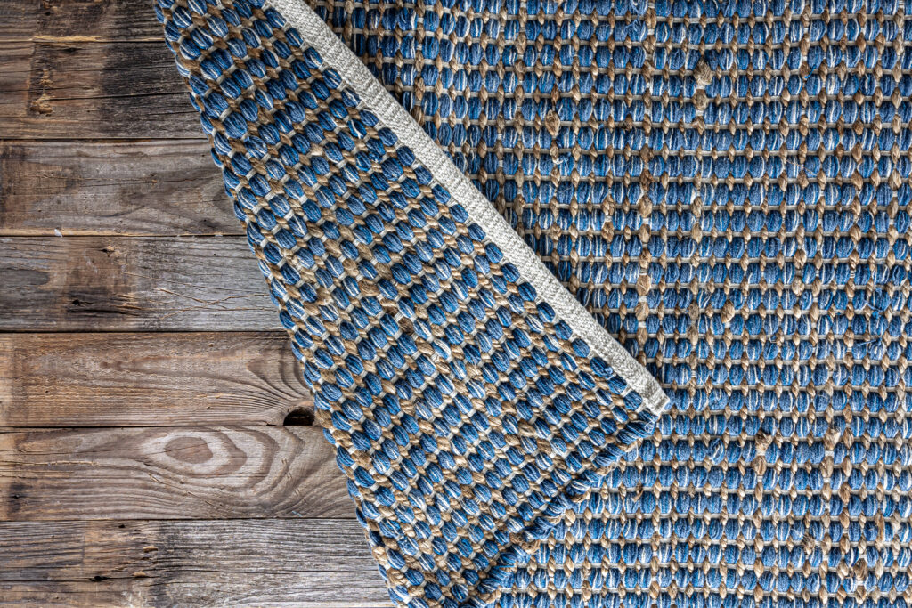 Tapis tressé en jute naturel et fibres de denim bleu posé sur un sol en bois vieilli. Tissage géométrique fait main idéal pour une décoration style industriel ou bord de mer.